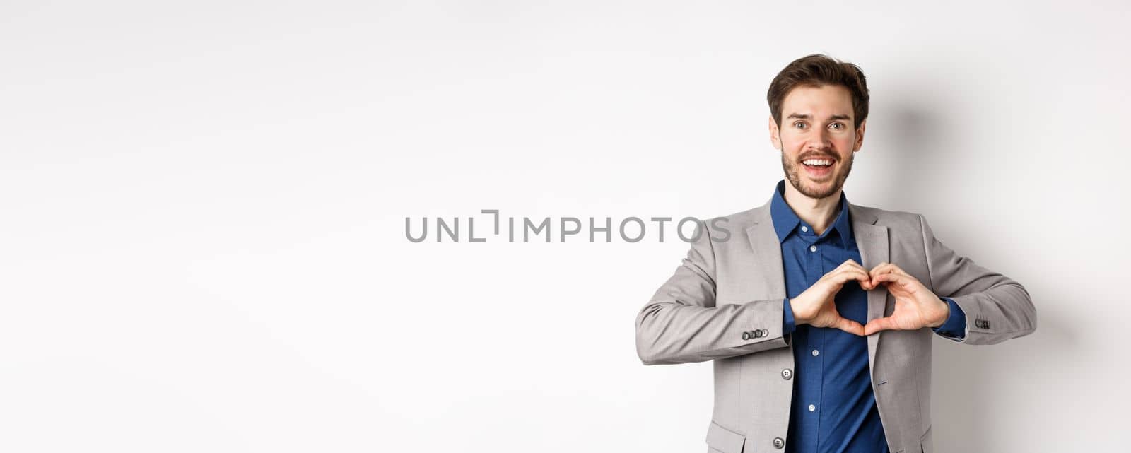 Passionate and romantic man in suit falling in love, showing heart sign and smiling at lover, standing on white background.