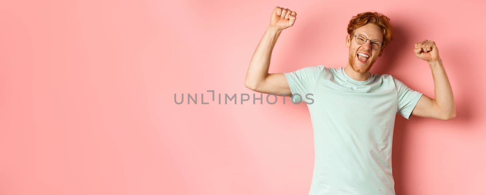 Cheerful young man with red hair looking happy, raising hands up in fist pumps gesture, celebrating success, feel like champion, winning and standing over pink background.