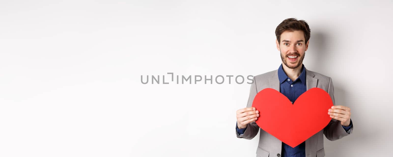 Happy man confessing in love, showing heart cutout and smiling at camera, standing in suit on romantic date with lover, white background.