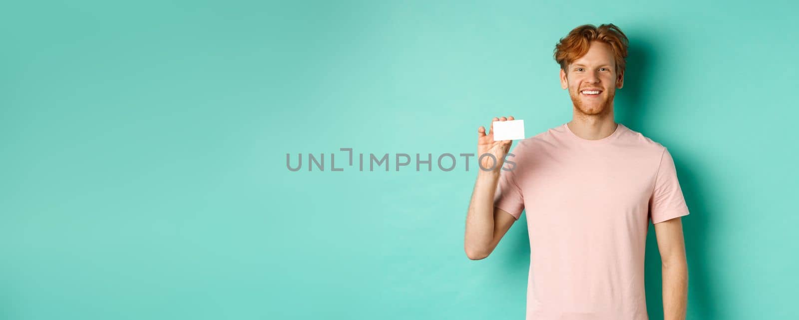 Handsome young man smiling and showing plastic credit card, standing in t-shirt against turquoise background by Benzoix