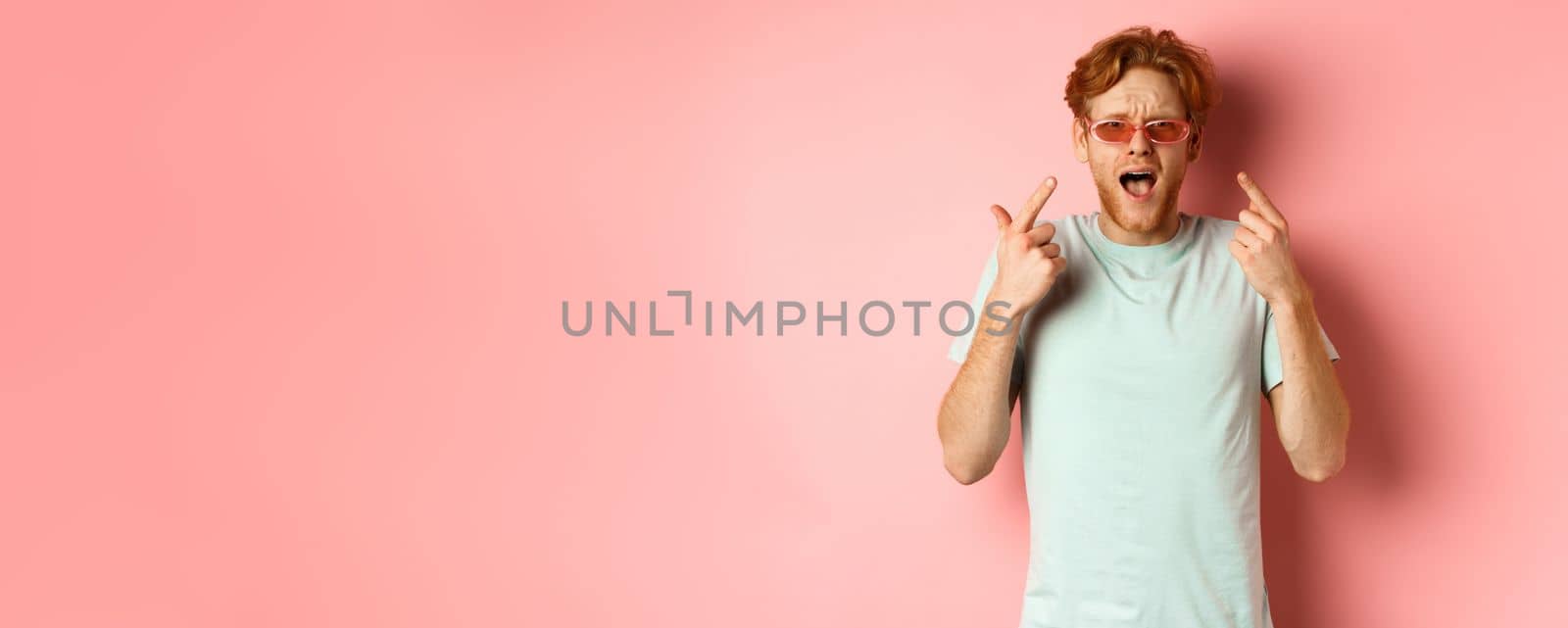 Young handsome man with red hair and beard pointing fingers at sunglasses, showing new glasses, standing over pink background.