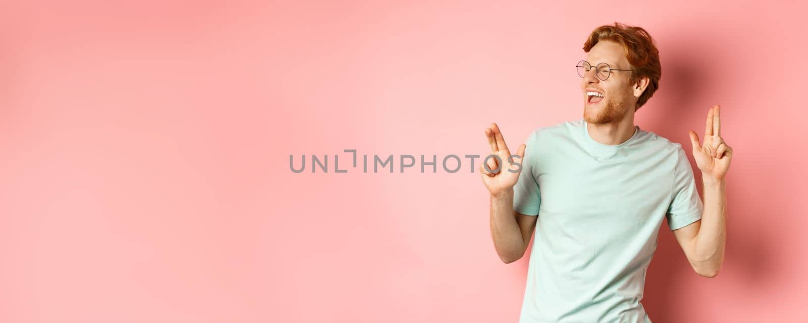 Cheerful redhead guy listening music and dancing, triumphing or celebrating success, standing over pink background by Benzoix
