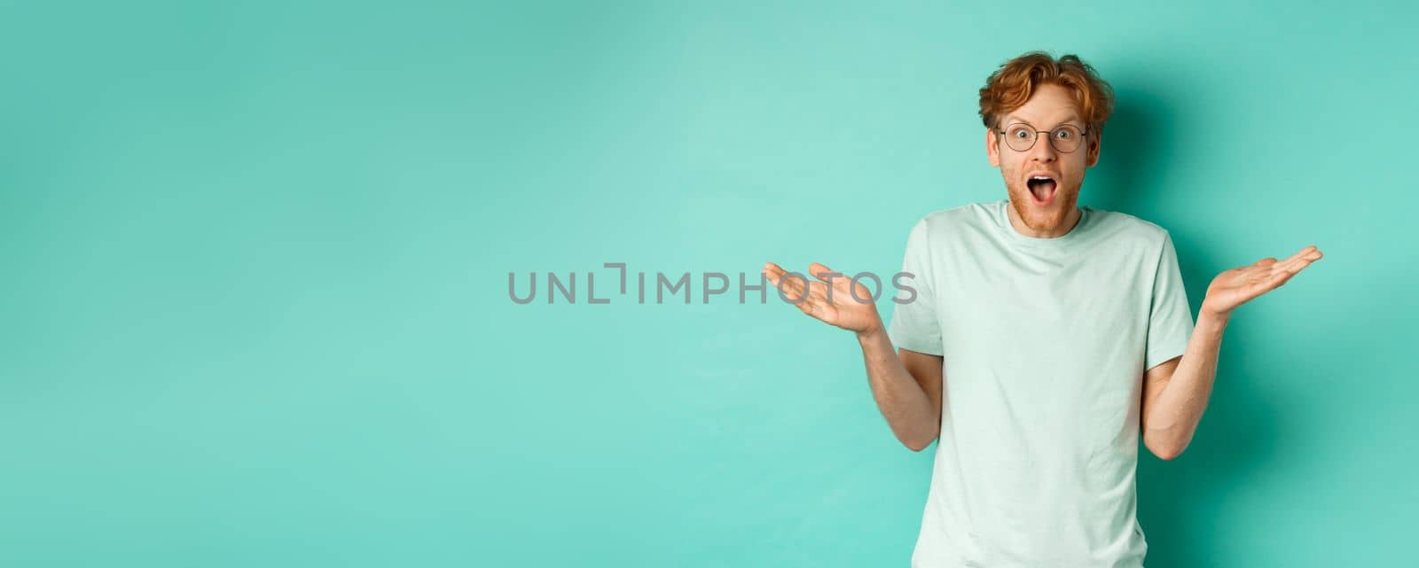 Surprised handsome man with red hair, wearing glasses, spread hands sideways and gasping in awe, looking wondered at camera, standing over turquoise background.