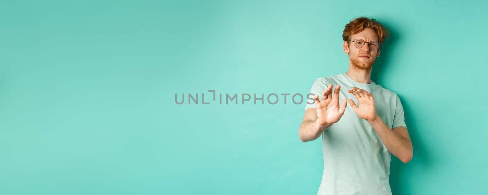 Reluctant redhead man in glasses raising hands in refusal gesture, step backwards, avoiding something with disgusted and unamused face, standing over turquoise background by Benzoix