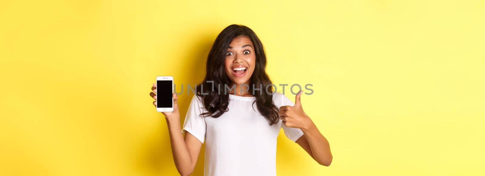 Portrait of excited african-american girl, showing smartphone screen and making thumbs-up, recommending something cool, standing over yellow background.