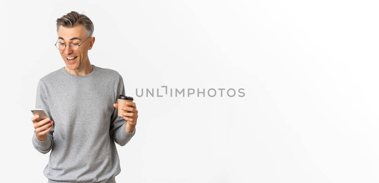 Image of handsome middle-aged man in grey sweater and glasses, drinking coffee and looking happy at smartphone screen, reading something online, standing over white background.