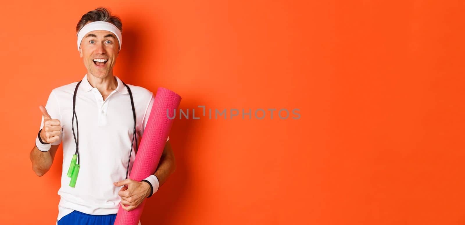 Concept of workout, gym and lifestyle. Cheerful middle-aged fitness guy, holding skipping rope and yoga mat, showing thumbs-up and smiling satisfied, standing over orange background.