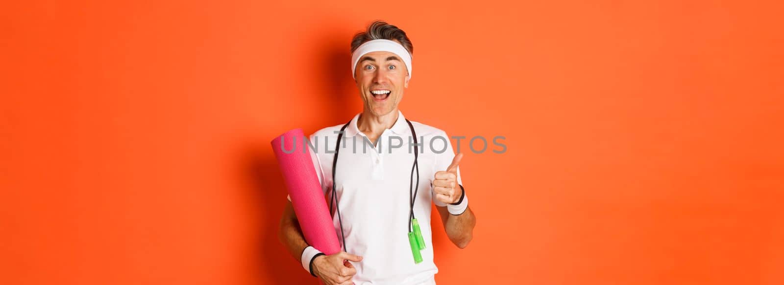 Concept of workout, gym and lifestyle. Cheerful middle-aged fitness guy, holding skipping rope and yoga mat, showing thumbs-up and smiling satisfied, standing over orange background.