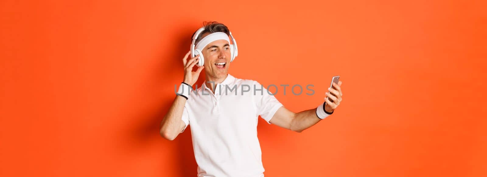 Portrait of handsome middle-aged male athlete, wearing gym uniform, listening music in headphones and taking selfie on mobile phone during workout, standing over orange background.