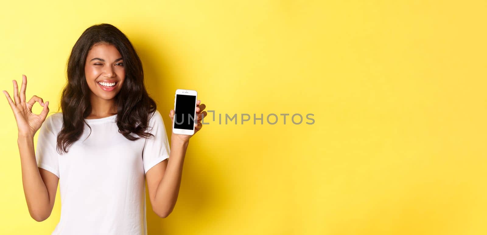 Image of beautiful african-american girl, winking and smiling, showing smartphone screen and okay sign in approval, standing over yellow background.