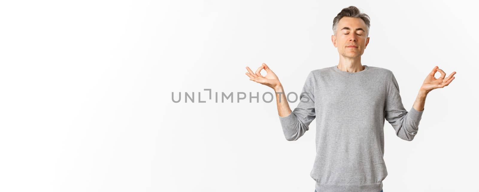 Portrait of calm and relaxed middle-aged man, breathing air freely, meditating with eyes closed and hands spread sideways, practice yoga over white background.