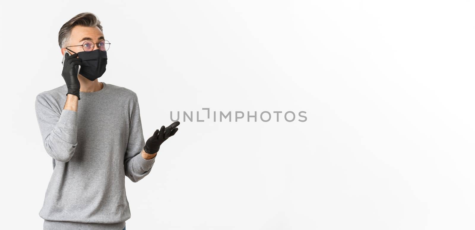 Concept of covid-19, social distancing and lifestyle. Image of handsome middle-aged man in medical mask and gloves, talking on mobile phone, standing over white background.