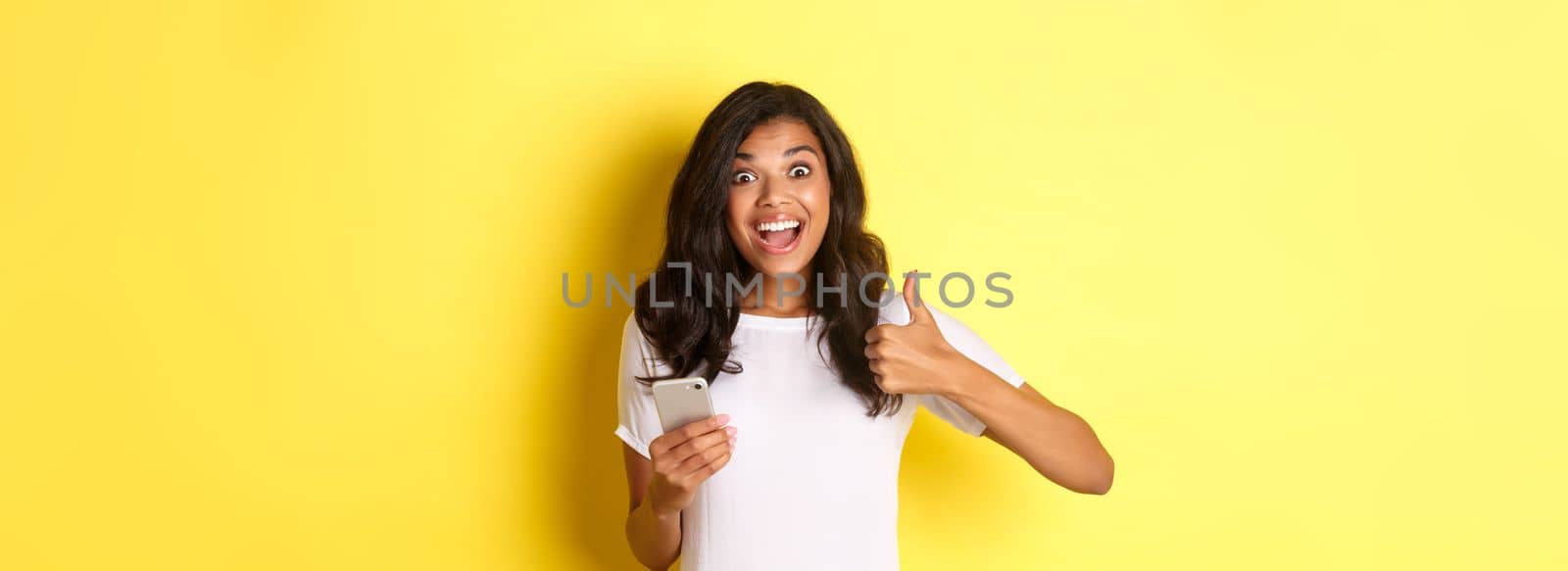Portrait of amazed african-american girl, showing thumbs-up and using mobile phone, praising cool app, standing over yellow background by Benzoix
