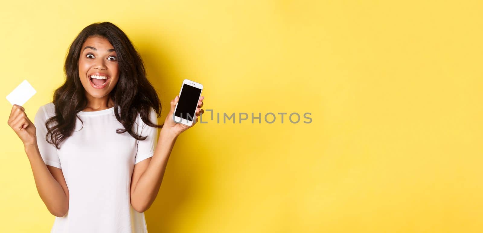 Image of excited african-american female model, showing smartphone screen and credit card, standing over yellow background.