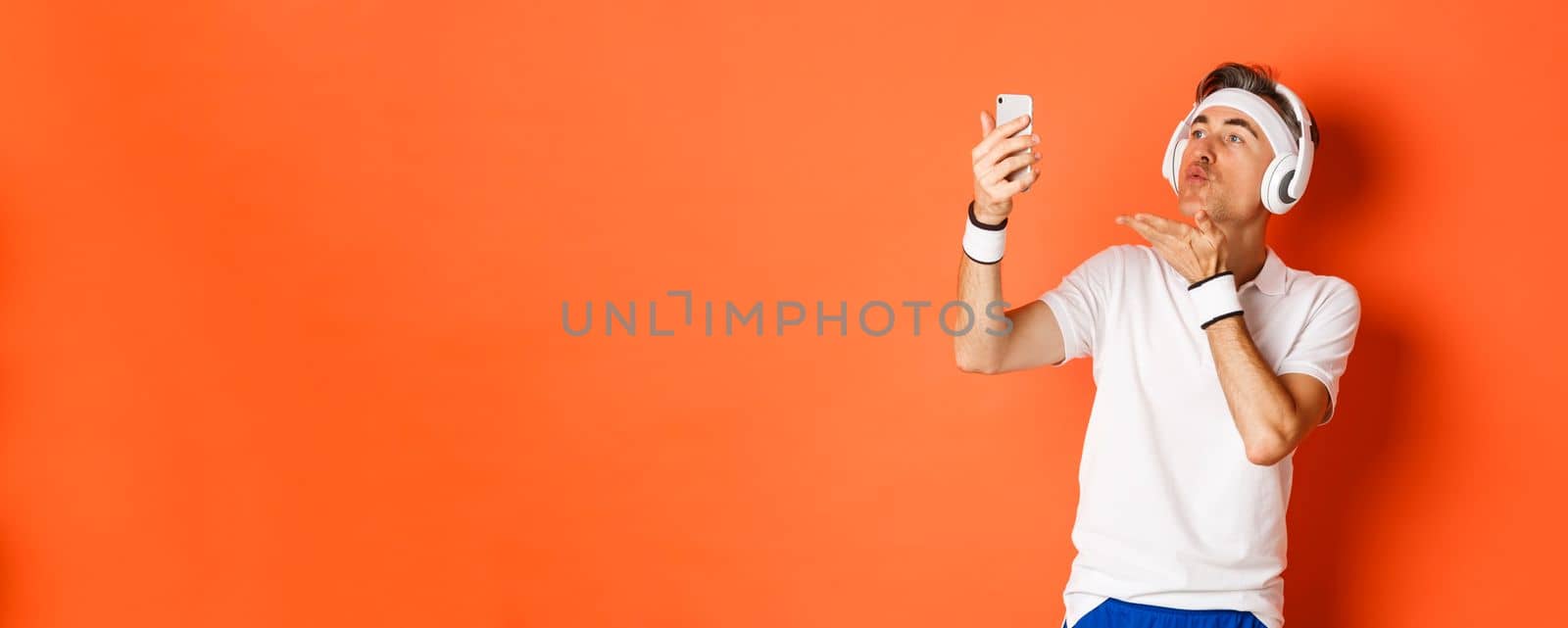 Portrait of attractive middle-aged man in gym uniform, wearing headphones, sending air kiss at mobile phone camera, taking selfie or having video call, standing over orange background by Benzoix