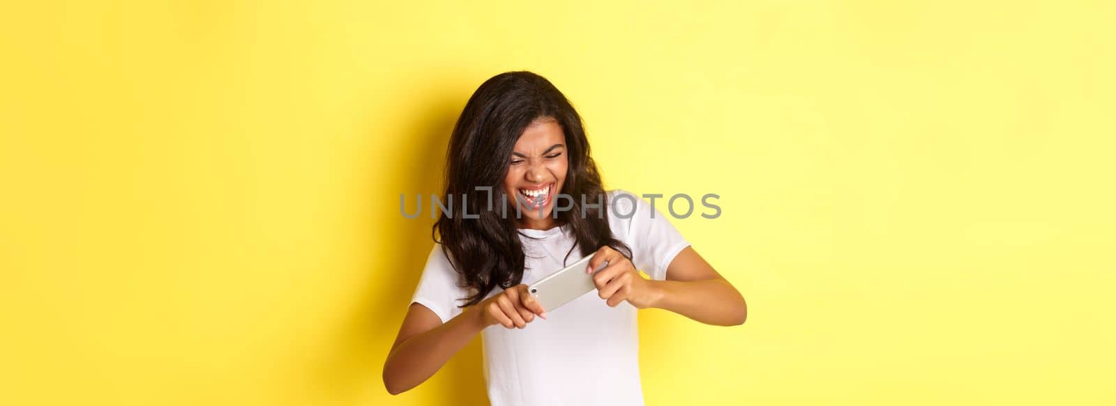 Portrait of cheerful african-american girl, playing mobile game and looking happy, standing over yellow background by Benzoix