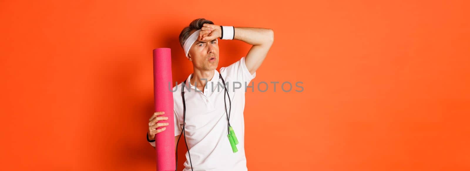 Concept of workout, gym and lifestyle. Image of handsome middle-aged man, tired after fitness exercises, holding skipping rope and yoga mat, wiping sweat off forehead, orange background.