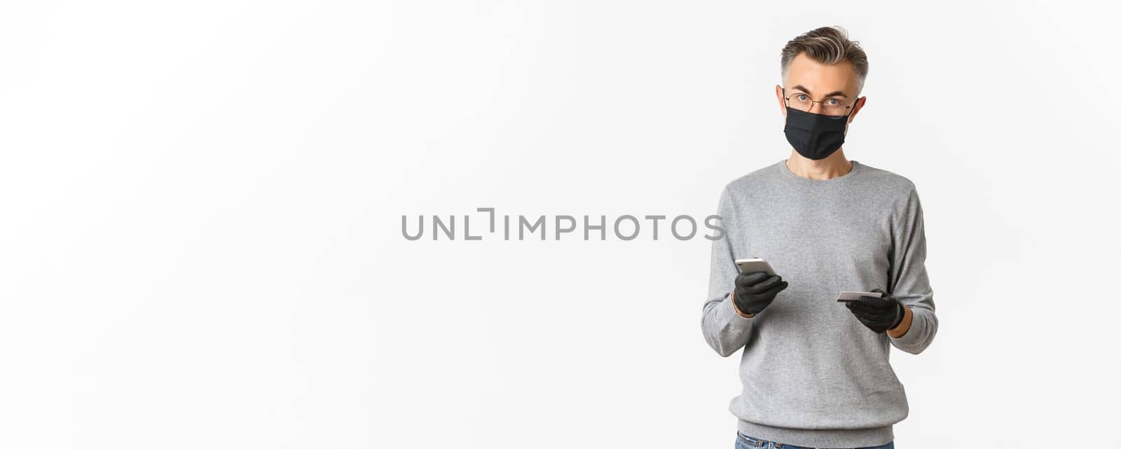 Concept of covid-19, social distancing and lifestyle. Attractive middle-aged guy in medical mask, gloves and glasses, shopping online with smartphone and credit card, standing over white background by Benzoix