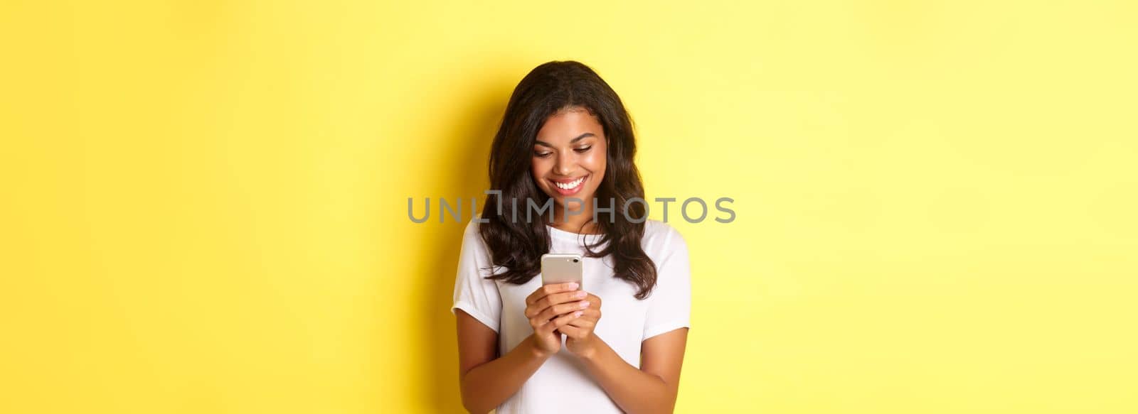 Portrait of good-looking african american girl in white t-shirt, messaging with smartphone, using mobile phone app, standing over yellow background by Benzoix