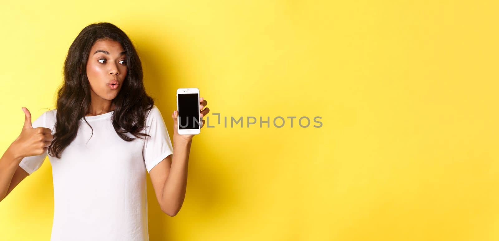 Portrait of good-looking african american female model, recommending an app, showing mobile phone and look excited, standing over yellow background.