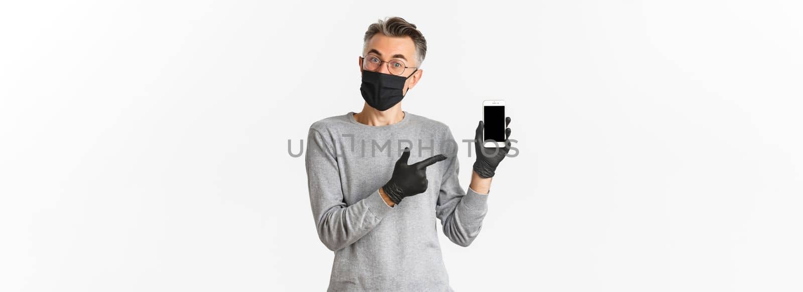 Concept of covid-19, social distancing and lifestyle. Image of attractive middle-aged guy in glasses, medical mask and gloves, explain something and pointing at mobile phone screen by Benzoix