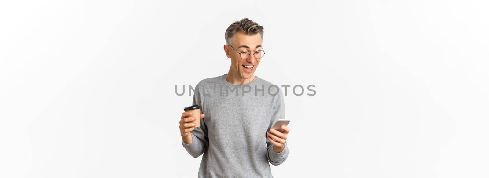 Image of handsome middle-aged man in grey sweater and glasses, drinking coffee and looking happy at smartphone screen, reading something online, standing over white background by Benzoix