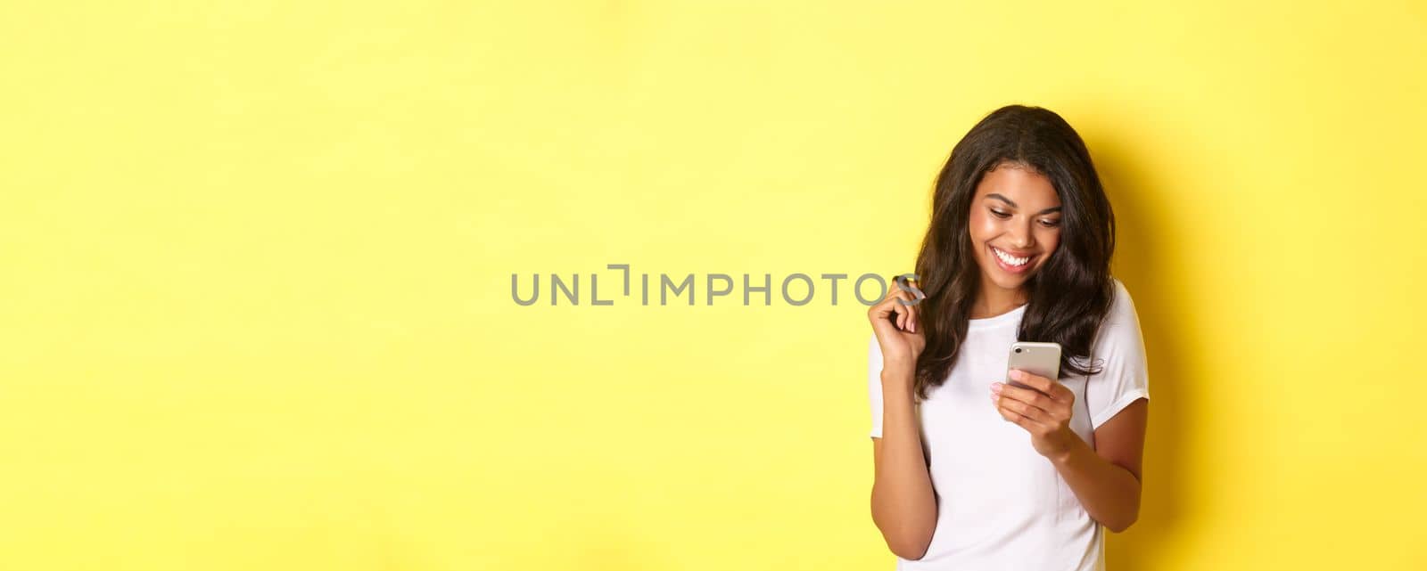 Image of attractive african american girl in white t-shirt, messaging on smartphone, looking at mobile phone and smiling, standing over yellow background.
