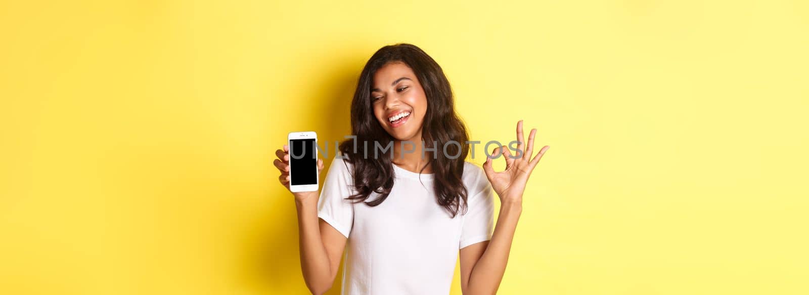 Portrait of cute african-american girl smiling pleased, showing okay sign and mobile phone screen, recommending an app or promo, standing over yellow background.