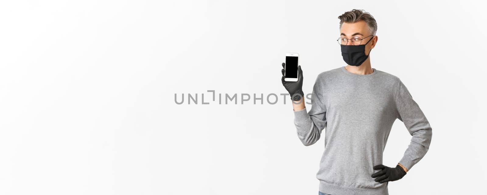 Concept of covid-19, social distancing and lifestyle. Portrait of handsome middle-aged man in medical mask, gloves and glasses, showing mobile phone screen, standing over white background.