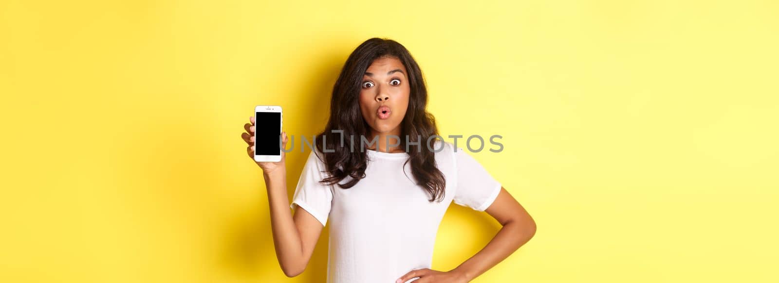 Image of amazed african-american girl, looking fascinated and showing smartphone screen, standing over yellow background by Benzoix