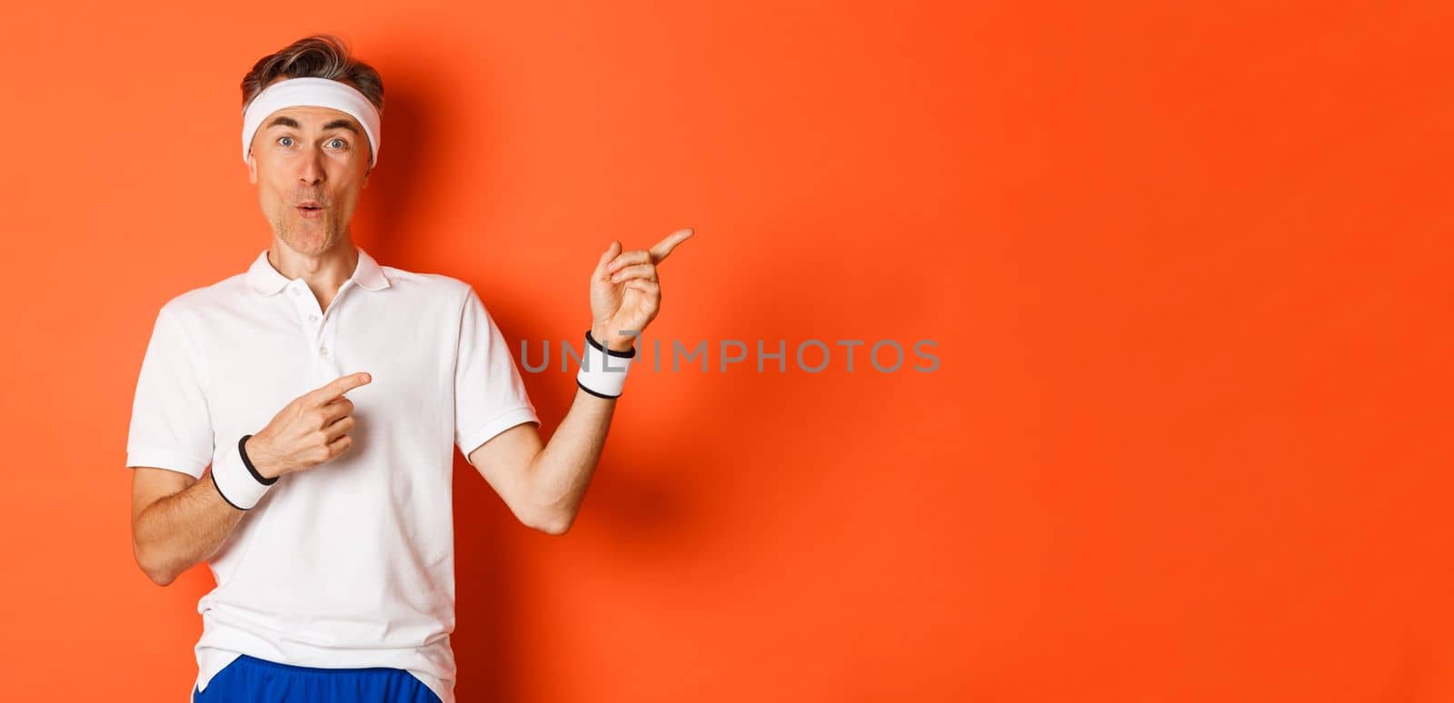 Concept of workout, sports and lifestyle. Portrait of amused middle-aged male athlete, pointing fingers at upper right corner, showing cool promo offer, standing over orange background.