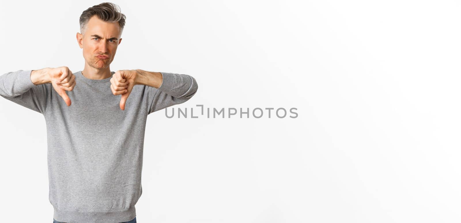 Skeptical and disappointed middle-aged man, grimacing unamused and showing thumbs-down, dislike something bad, standing over white background.