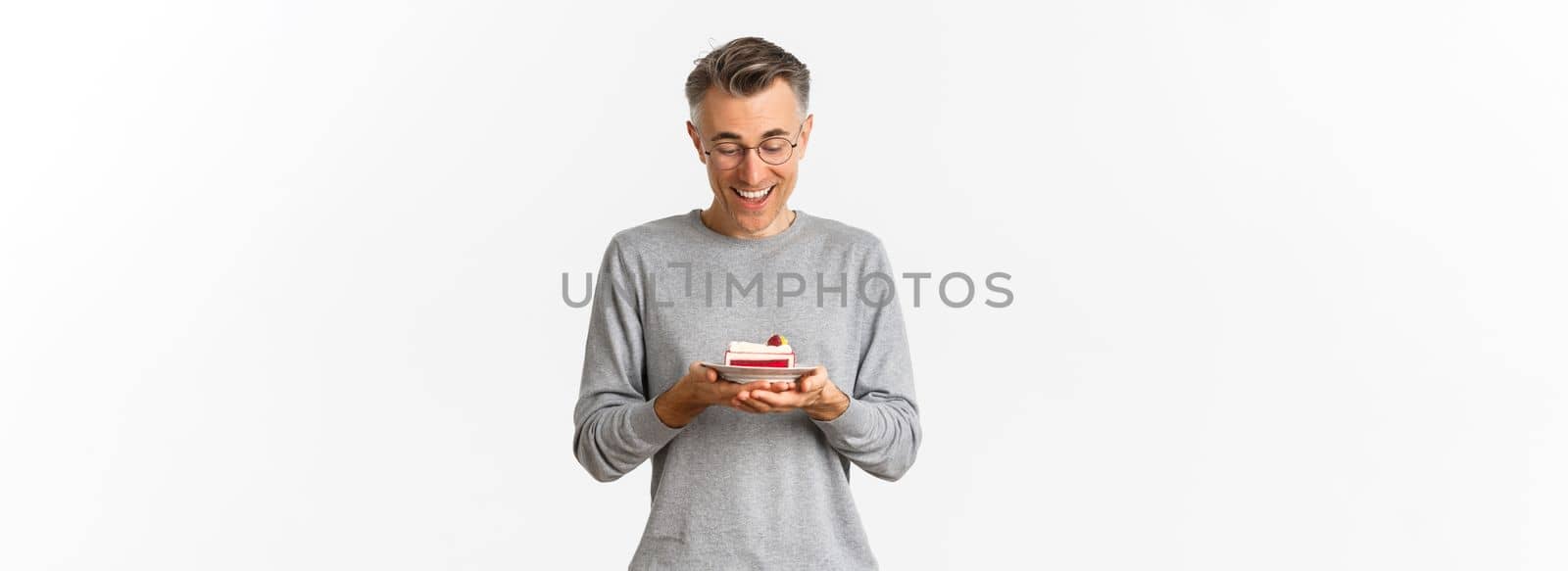 Portrait of handsome middle-aged man, looking happy at delicious cake, standing over white background.