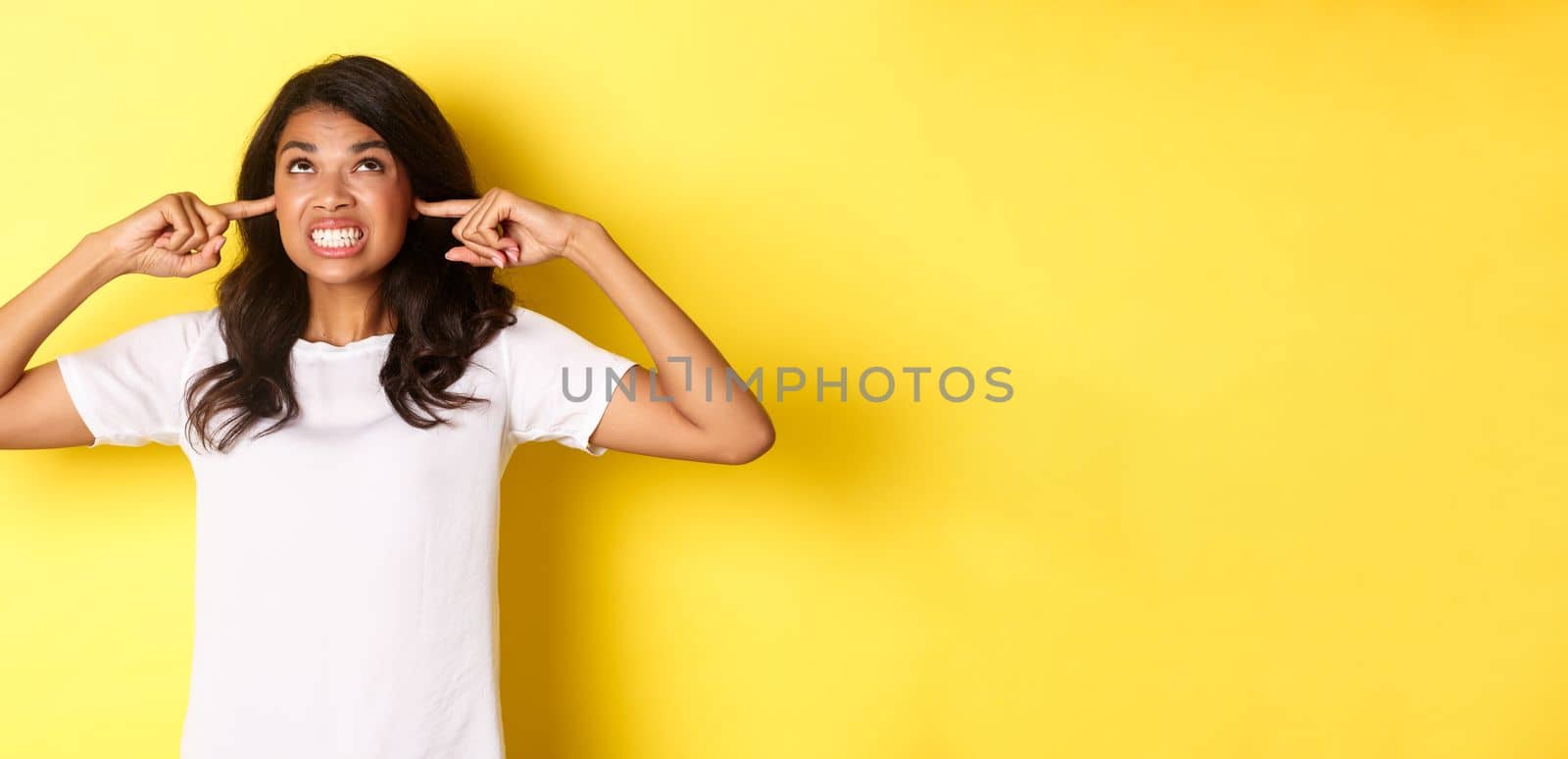 Image of annoyed and bothered african-american girl, shut ears and looking angry up, complaining at loud neighbours, standing over yellow background.