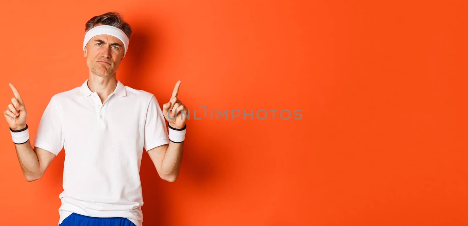 Portrait of skeptical adult sportsman in headband and t-shirt, complaining and pointing fingers up, looking disappointed, standing over orange background.
