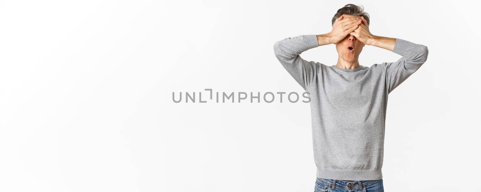 Image of excited middle-aged guy in grey sweater, cover eyes with hands and looking amazed while waiting for surprise, playing hide-n-seek, standing over white background.