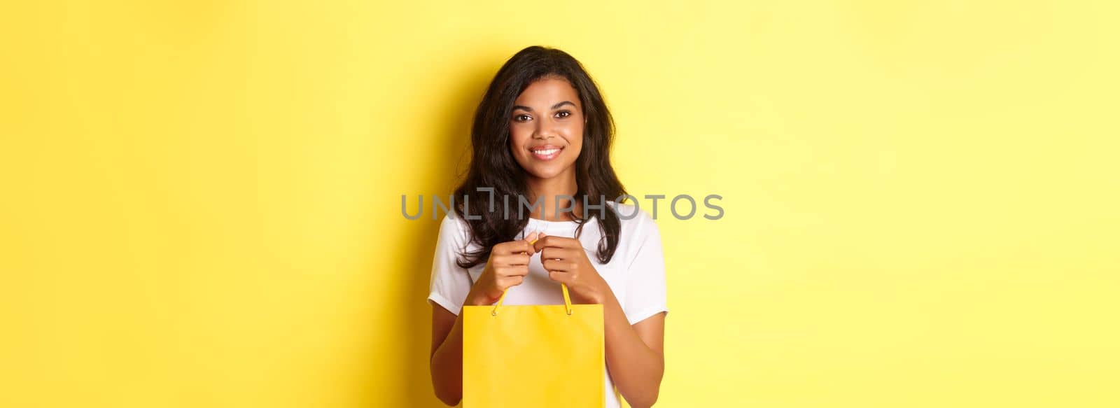 Image of attractive african-american woman, holding shopping baf and smiling, standing over yellow background.