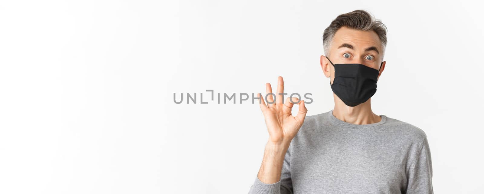 Concept of covid-19, social distancing and quarantine. Close-up of surprised and happy middle-aged man, wearing black medical mask, showing okay sign and look amazed, white background.