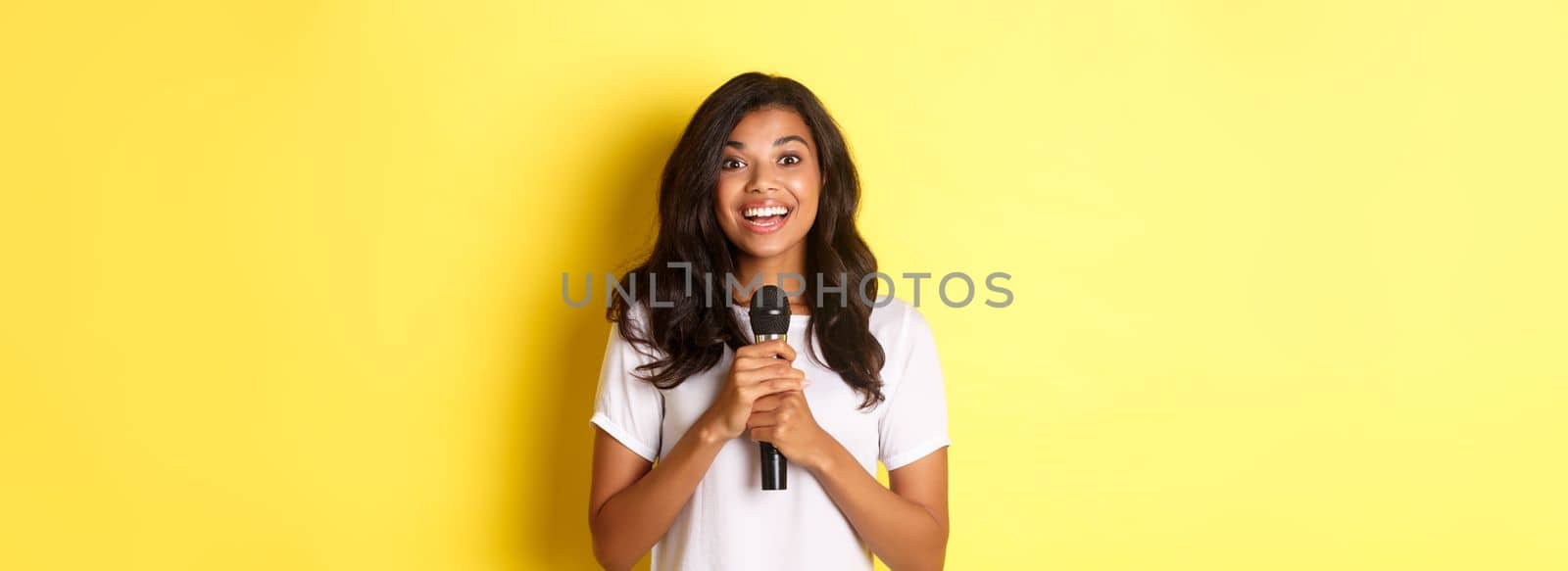 Portrait of happy african-american girl, looking amused while giving speech, holding microphone and smiling at camera, standing over yellow background.