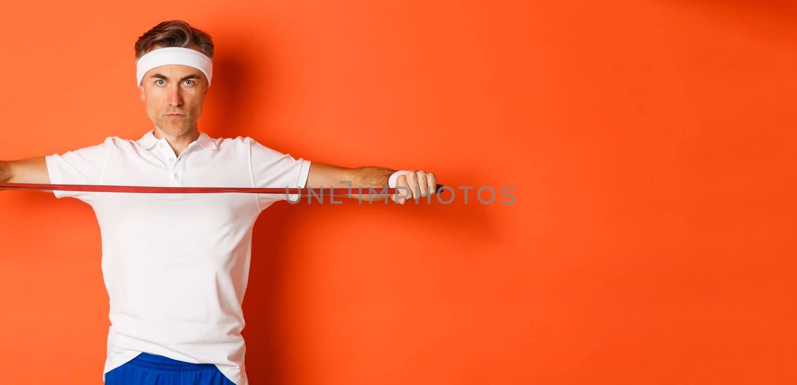 Concept of workout, gym and lifestyle. Serious and confident middle-aged fitness guy, holding stretching rope for exercises, standing over orange background by Benzoix