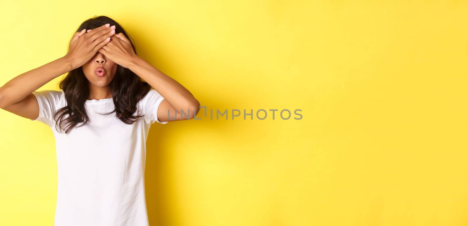 Image of excited african-american girl waiting for surprise, smiling and covering eyes with hands, standing over yellow background.