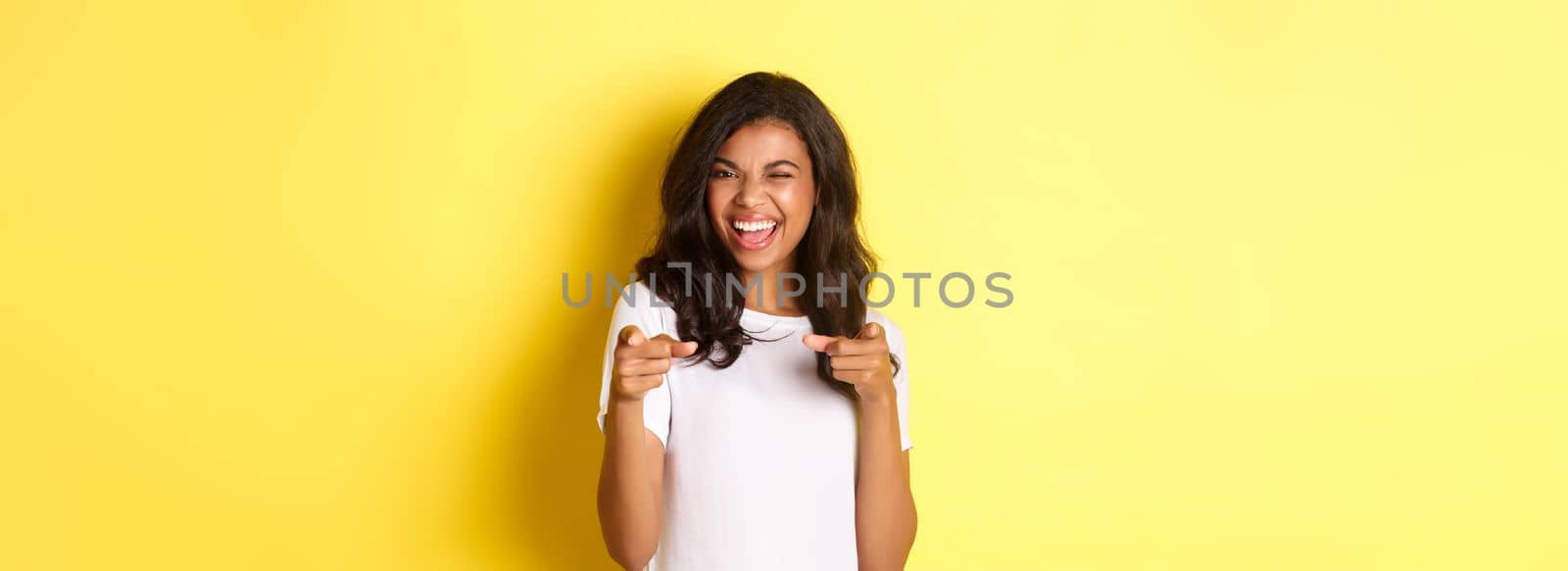 Portrait of cheeky, beautiful african-american woman in white t-shirt, winking and pointing fingers at camera to say congrats, praising you, standing over yellow background.