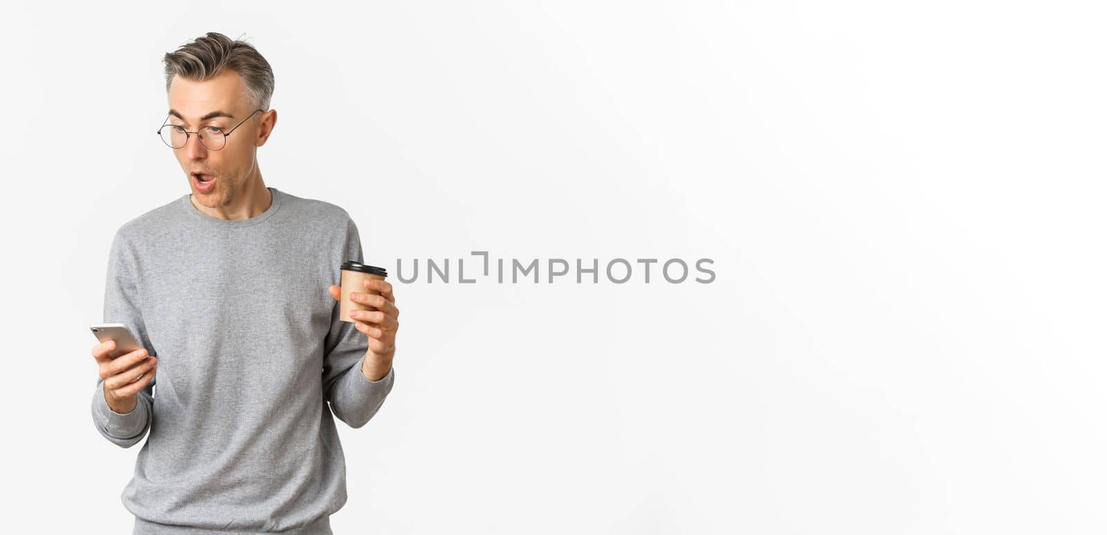 Image of handsome middle-aged man in grey sweater and glasses, drinking coffee and looking amazed at mobile phone screen, reading something online, standing over white background by Benzoix
