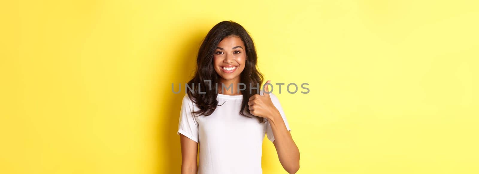 Portrait of happy and cute african-american girl, smiling pleased and showing thumbs-up in approval, like and approve something, standing over yellow background by Benzoix