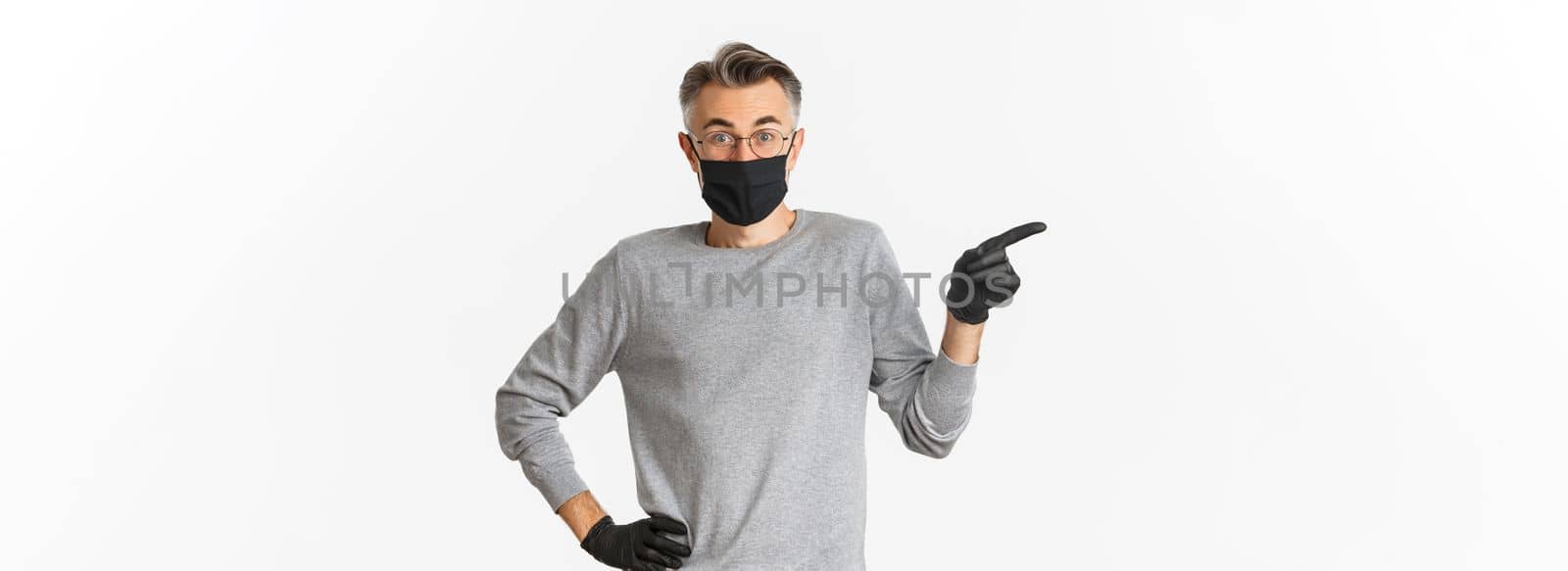 Concept of coronavirus, lifestyle and quarantine. Portrait of handsome middle-aged man in glasses, medical mask and gloves, asking question with curious face, pointing finger left, white background.