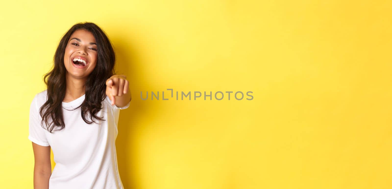 Portrait of beautiful and happy african-american woman laughing, pointing finger at you, found excellent candidate, standing over yellow background.