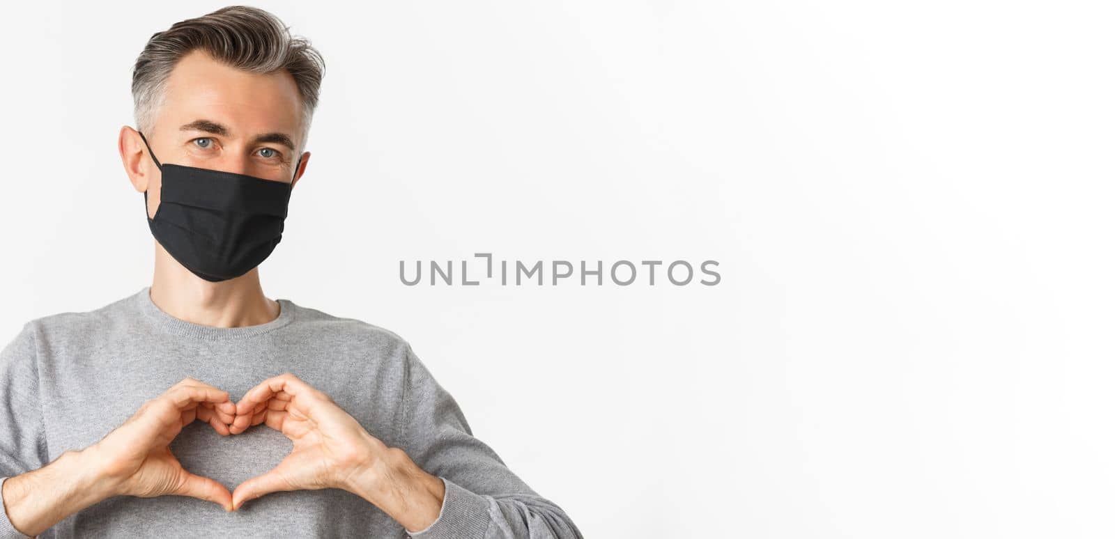 Concept of covid-19, social distancing and quarantine. Close-up of attractive middle-aged man in black medical mask, showing heart sign and smiling, standing over white background.