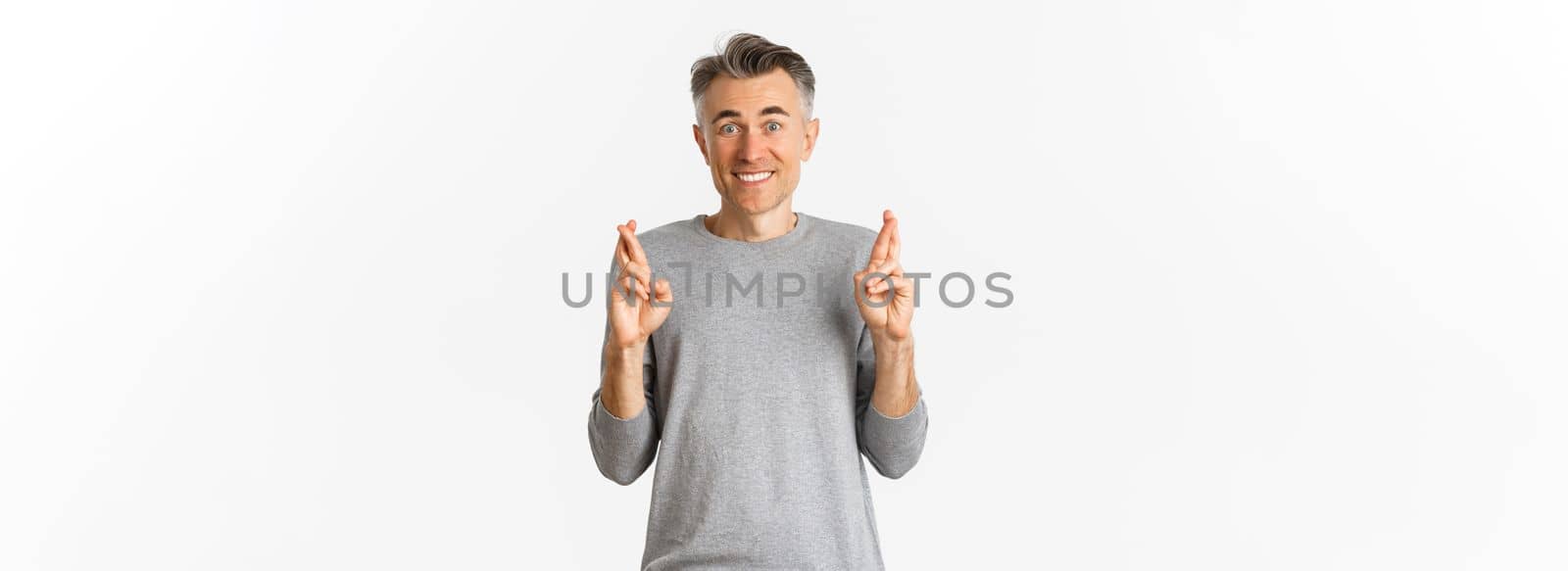 Image of excited and hopeful middle-aged man making a wish, waiting for results, cross fingers for good luck and smiling, standing over white background.