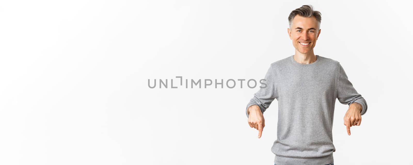 Handsome and confident middle-aged man in grey sweater, pointing fingers down and smiling, showing good deal promo, standing over white background.