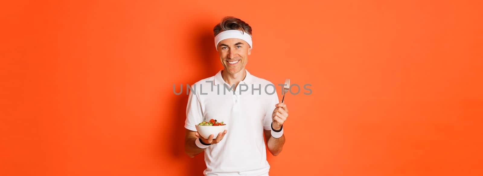 Concept of sport, fitness and lifestyle. Image of handsome, healthy and active male athlete, eating salad and smiling, standing over orange background.